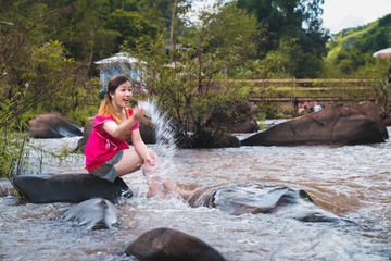 A beautiful woman wearing a pink dress sitting on a rock in a stream and splashing water with her hands In the midst of nature surrounded by mountains and forests