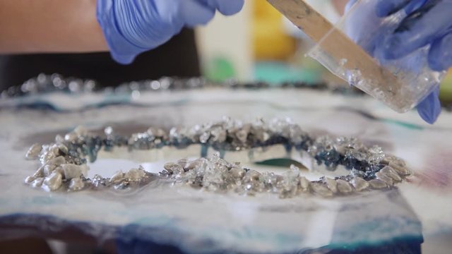 Closeup of hands of artist in protective gloves using wooden stick to decorate unfinished geode style painting with glitter and sparkling stones