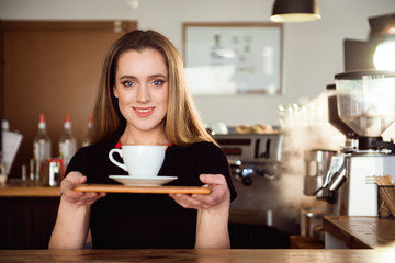 Beautiful female barista is working in coffee shop. Attractive woman is standing behind the bar counter, making coffee and welcomes customers.