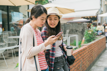 Image of cheerful asian korean women travelers in casual clothes drinking takeaway coffee and using smartphone on red brick wall outdoors. two ladies enjoy sunshine sharing mobile phone together.