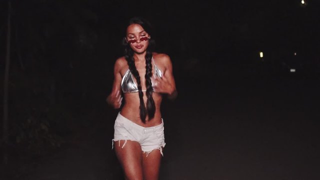 Cute and sexy brunette girl in silver bikini top, denim shorts and sunglasses dancing, smiling, flirting during summer evening - video in slow motion