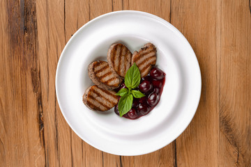 Grilled meat pieces with berry sauce served on a white plate over dark rustic wooden background. Italian cuisine.