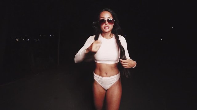 Cute and sexy brunette girl wearing pink long sleeve crop top and panties set and sunglasses dancing, smiling, flirting during summer evening - video in slow motion