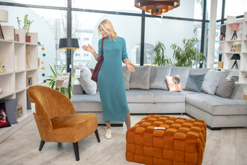 Blond-haired woman standing in indecision in a furniture store. - obrazy, fototapety, plakaty