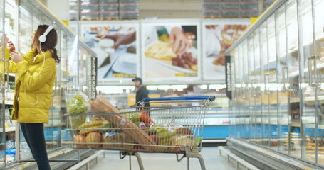 Joyful Caucasian young girl in headphones choosing products in supermarket. Cheerful woman...