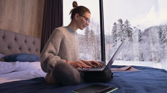 Portrait of attractive freelancer woman with glasses and a sweater with stockings sitting on a bed in an eco-house in the middle of a winter forest with a laptop