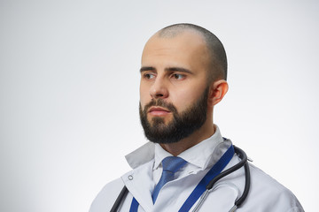 A close portrait of a doctor with a beard who looks away. A physician practitioner in a white lab coat, a blue tie, and a stethoscope. 