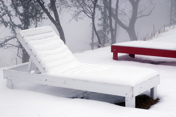 Wooden white deck chairs in snow. Snow covered sun loungers. Unexpected snow concept.