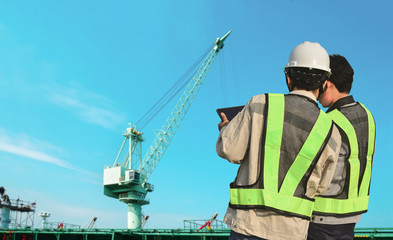 Male worker at construction site with hand holding iPad work wearing safety helmet hard hat on green pipe line and crane in project site background.