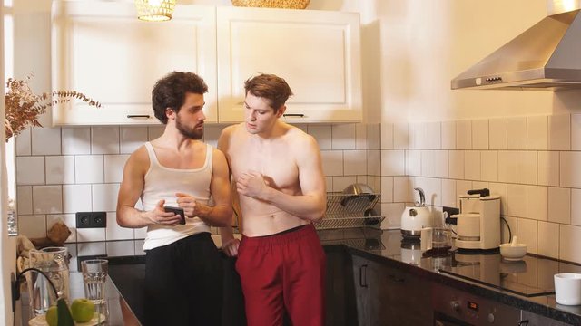 Morning, breakfast. Two young sexy guys in the kitchen. View the news feed of instagram using smartphone.