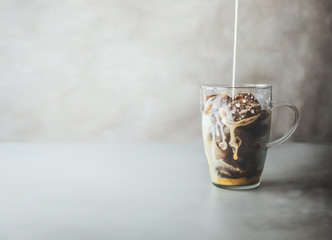 Ice coffee preparation. Cream milk pouring  in glass with coffee ice cubes in glass mug on rustic table at concrete wall background. Iced coffee making. Summer refreshing beverage. Cold drink.