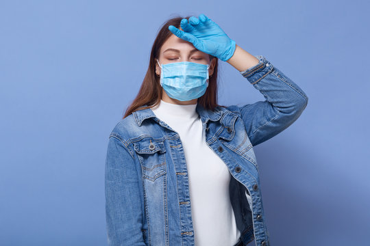 Image of exhausted sick young female having fever, putting one hand on forehead, feeling unwell, closing both eyes, having Covid19, wearing antibacterial mask and gloves. Coronavirus concept.