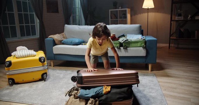 Funny little asian kid with curly hair is hurryingly packing a suitcase, filling it up with clothes, jumping on it to close it - travelling concept 4k footage