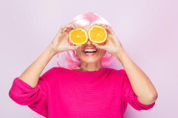 Caucasian woman with pink wig and sweater, with two orange slices in his eyes posing funny at the photo studio. Funny expressive women and colorful portrait concept. Pink and orange.