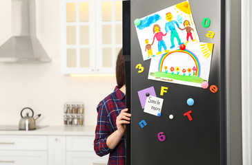 Woman opening refrigerator door with child's drawings, notes and magnets in kitchen. Space for text