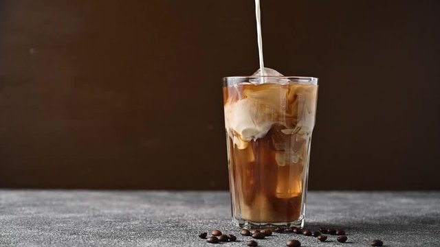 Add cream to iced coffee. Slow motion milk pouring into a glass cup with coffee and ice.