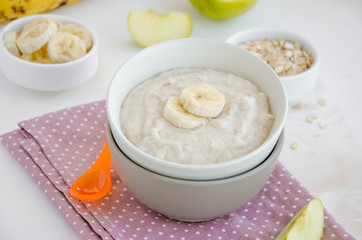 Baby food. Creamy oatmeal with banana slices and apple in a bowl with a spoon on a light background. Healthy breakfast. Porridge for breakfast. Horizontal orientation. Close up.