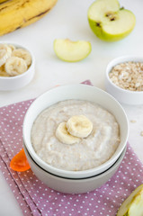 Baby food. Creamy oatmeal with banana slices and apple in a bowl with a spoon on a light background. Healthy breakfast. Porridge for breakfast. Vertical orientation. Copy space