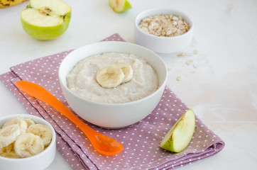 Baby food. Creamy oatmeal with banana slices and apple in a bowl with a spoon on a light background. Healthy breakfast. Porridge for breakfast. Horizontal orientation.