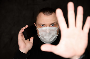 Photo portrait of an angry adult man in a mask with a coronavirus on a black background with a phone in his hands, shows his fingers