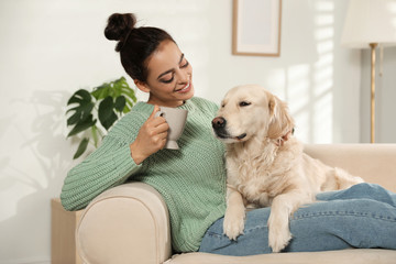 Young woman with cup of drink and her Golden Retriever on sofa at home. Adorable pet