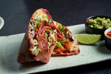 Close up view on burrito with salami chorizo, rice, tomatoes, corn and bell pepper in brown pita on a brown plate with tomato salsa and guacamole. Vegetarian Shawarma Roll