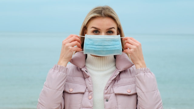 Close-up portrait of blonde woman putting medical mask on her face while quarantining virus Coronavirus COVID-19 stands on seashore and blue sky. Epidemic, pneumonia, hygiene, pandemic. Walk on aiк