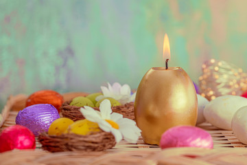 Obraz premium Greeting card Easter burning golden candle and chocolate eggs in colored foil. Blurred background and glowing bokeh. Copy space.