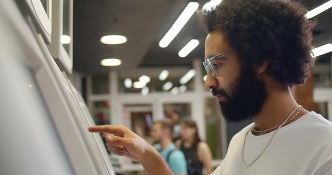 Close up view of man in glasses using ATM machine. Curly haired guy in 30s pressing on display while standing near self service terminal. Indoors. Concept of people and technology.