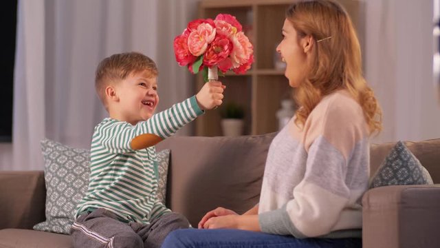 mother's day, holidays and family concept - happy little son giving flowers, kissing and hugging his mother at home in evening