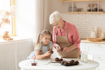 Lovely girl and her grandma with fresh chocolate muffins indoors, space for text