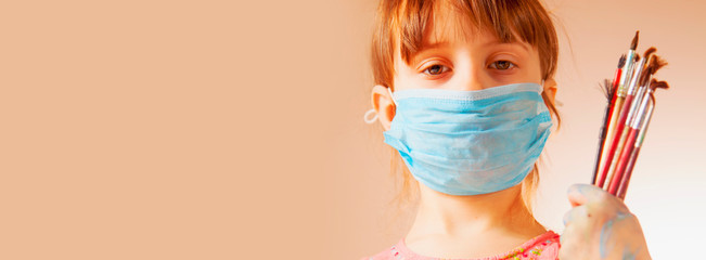 Close up portrait of cute little child girl in protect medical face maskholding paint brushes....