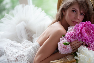 Bride in beautiful dress lying on sofa whit peonies in white studio interior. Romantic wedding style shot with sun light