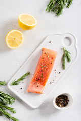 Fresh raw salmon fillet on white stone board with salt, peppers, lemon, and rosemary on white background, top view. Restaurant menu, recipe, keto diet, healthy nutrition