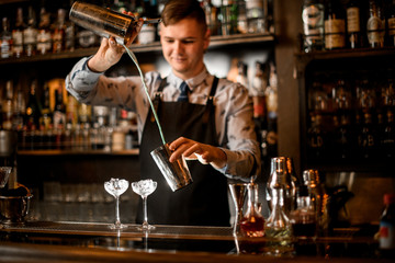 Barman pours cocktail in shaker. Two glasses with ice stand on bar.