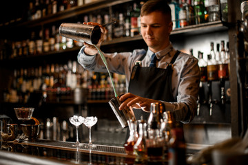 young bartender pours cocktail in shaker. Two glasses with ice stand on bar.