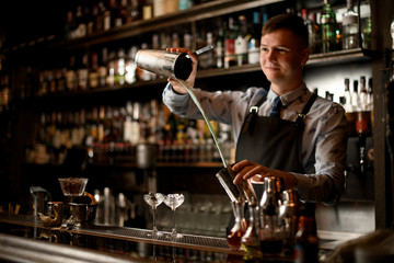 Young bartender professionally pours cocktail in shaker. Two glasses with ice stand on bar.