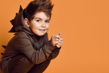 Little brunet male in brown dino hoodie with hood. He is smiling, posing sitting squatting against...