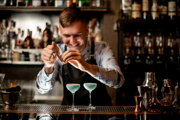 young smiling barman decorates two glasses with cocktails.