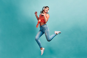 Young beautiful energy girl with white headphones listening to music laughs and jump on blue...