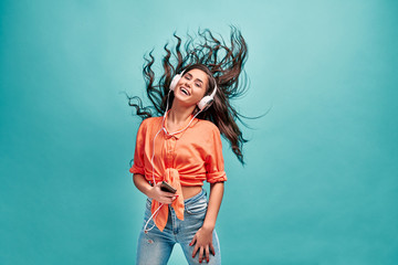 Beautiful energy girl with white headphones listening to music and laughs on blue background in studio. Long hair in is flying from moving.
