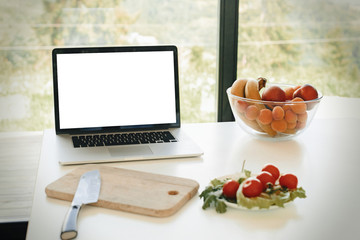 Laptop with blank white screen and vegetables, knife and fruits on countertop in modern kitchen with big windows, copy space. Culinary courses or recipe online. Cooking home concept. Food blog