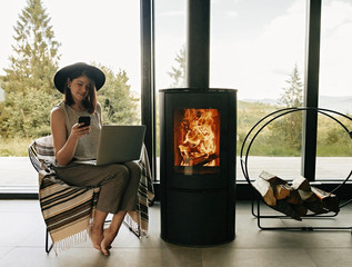 Stylish hipster girl with phone and laptop sitting on cozy chair near fireplace in modern living...