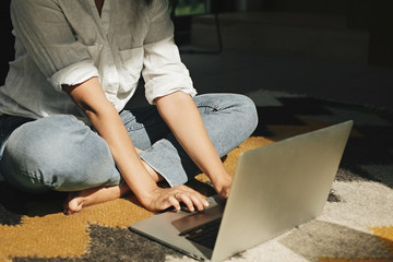 Stylish hipster girl using laptop, sitting on floor rug in modern room in sunny light. Young woman shopping or working online from home. Freelance and freelancer