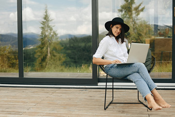 Young happy woman in hat using laptop, shopping or working online from home outside. Stylish hipster girl sitting with laptop on wooden porch at big window. Freelance and freelancer