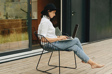 Stylish hipster girl sitting with laptop on wooden porch at big window. Young happy woman in hat...