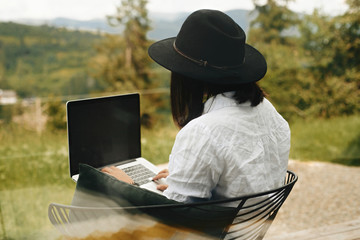 Hipster girl with laptop sitting on wooden porch with beautiful view on woods and mountains....