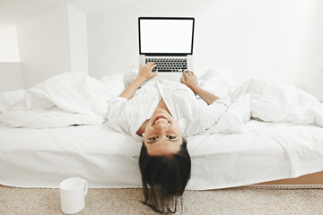 Happy stylish girl  lying on bed with laptop with blank screen and smiling in white modern room....