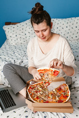 Woman works with a laptop in bed and eats a pizza delivery. Fast food, pizza concept.