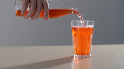 Hand pouring an orange carbonated drink from glass bottle into a glass. Male hand Holding orange soda bottle tight and close pours Into a glass Outside. Pouring fresh and cold yellow lemonade drink.